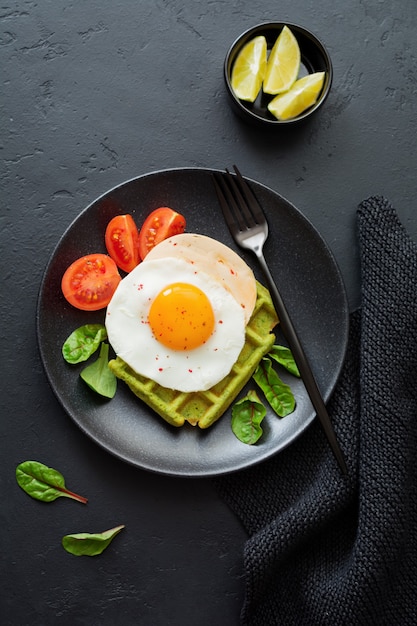 Œuf Frit Avec Du Fromage, Des Tomates, Des Blettes Et Des Gaufres Aux épinards Sur Une Plaque En Céramique Noire Sur Une Table En Béton Foncé. Sandwich Pour Le Petit Déjeuner. Mise Au Point Sélective. Vue De Dessus. Copiez L'espace.