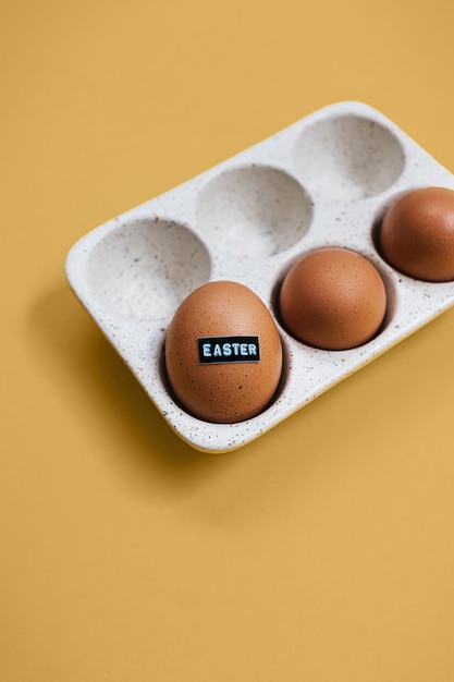 Oeuf de poulet cru brun avec mot en relief Pâques et oeufs dans un plateau de rangement en céramique sur fond jaune avec espace de copie Décor de vacances créatif et carte de voeux de printemps Mise à plat