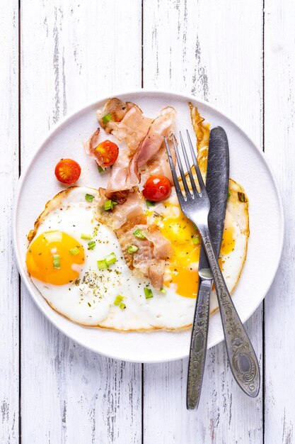 Oeufs au plat avec du bacon sur une plaque blanche et un fond en bois blanc.