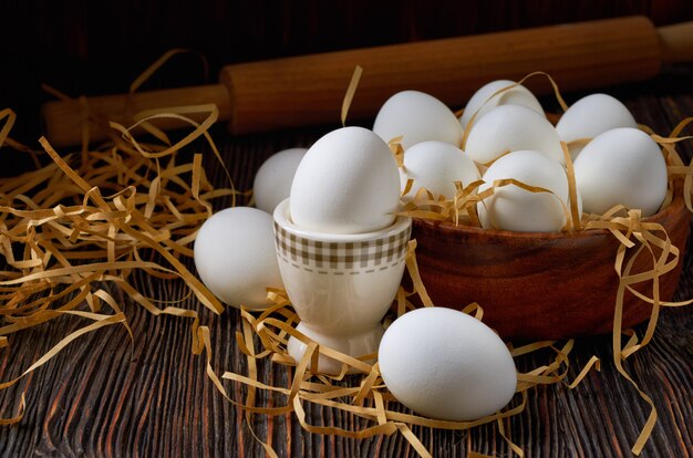 Oeufs blancs dans un bol en bois, sur une paille en papier et une table en bois. Au dos d'un rouleau à pâtisserie. Discret.