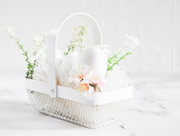 Oeufs blancs décoratifs dans un panier en métal avec des oeufs fleurs printanières et de la paille sur une table en marbre