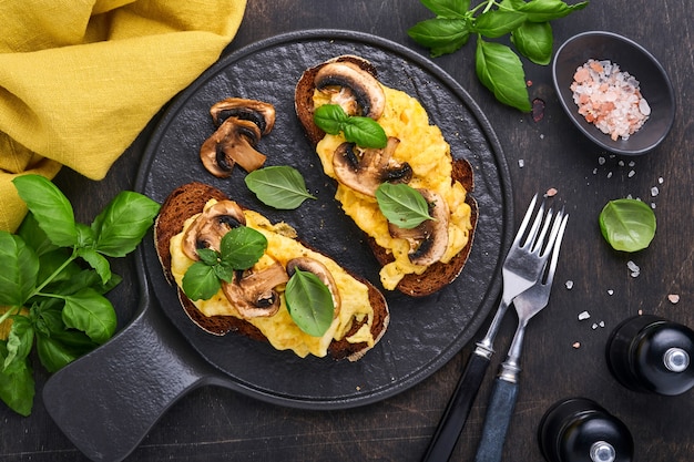 Oeufs brouillés aux champignons frits et basilic sur pain sur fond de tableau noir. Petit-déjeuner ou brunch fait maison - sandwichs aux œufs brouillés et aux champignons. Vue de dessus avec espace de copie