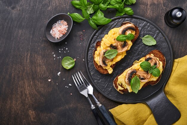 Oeufs brouillés aux champignons frits et basilic sur pain sur fond de tableau noir. Petit-déjeuner ou brunch fait maison - sandwichs aux œufs brouillés et aux champignons. Vue de dessus avec espace de copie