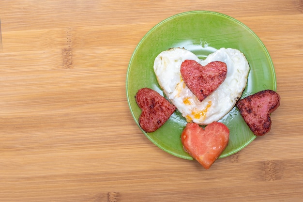 Oeufs brouillés en forme de coeur avec saucisses et tomate en forme de coeur sur une plaque verte sur fond de bambou, vue de dessus, espace de copie. Petit-déjeuner pour les proches le jour de la Saint-Valentin