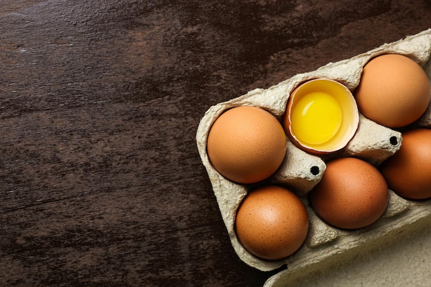 Oeufs bruns frais et un oeuf cassé avec du jaune dans un bac écologique en papier recyclé sur un bois sombre