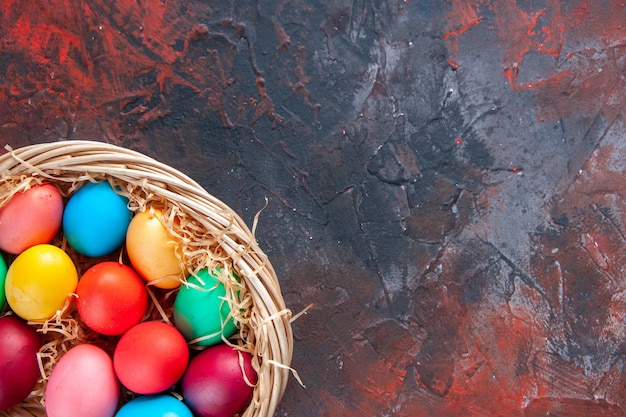 Oeufs colorés déposés côte à côte dans un panier de paille jaune sur le côté droit sur fond de couleurs sombres