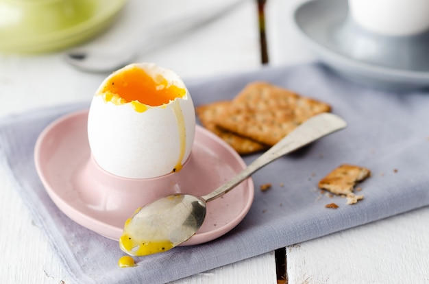 Oeufs à la coque dans des coquetiers avec cuillères et craquelins aux graines sur fond de bois blanc