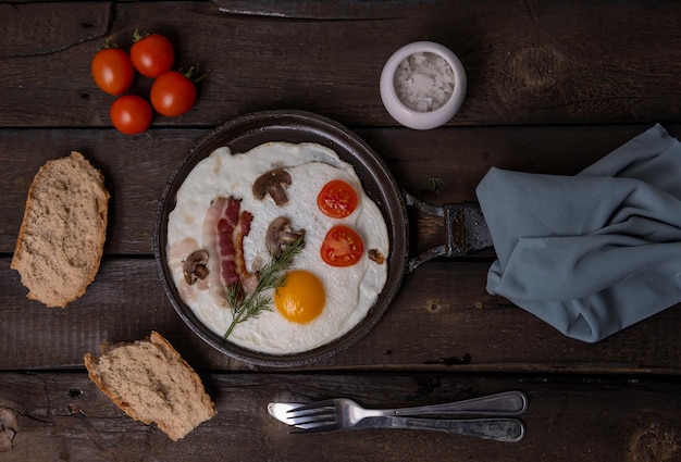 Des œufs frits avec du bacon et des légumes dans une poêle à frire en fonte sur un vieux fond de table en bois foncé