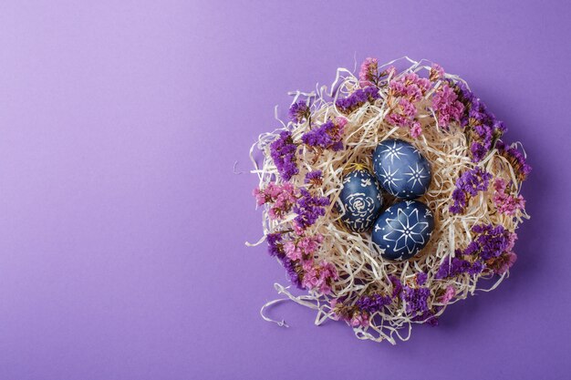 Oeufs de Pâques bleu foncé peints à la main dans le nid et fleurs sur fond violet