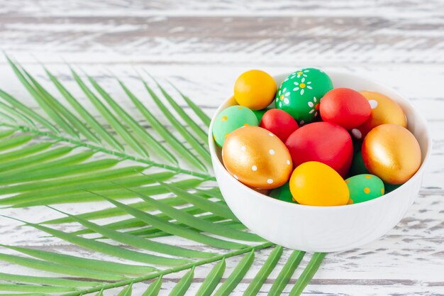 Oeufs de pâques colorés à la mode, feuilles tropicales vertes et plumes sur une assiette sur une table en bois. Préparation des vacances de Pâques. Composition festive