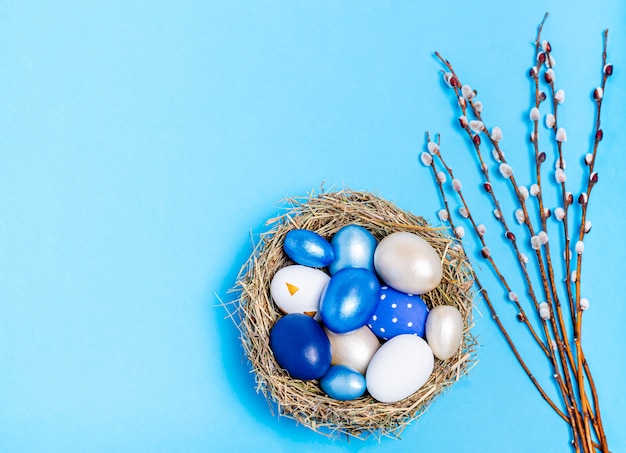 Oeufs de Pâques de fleurs bleues dans un nid décoratif de branches de paille et de saule sur fond bleu, espace copie, mise à plat