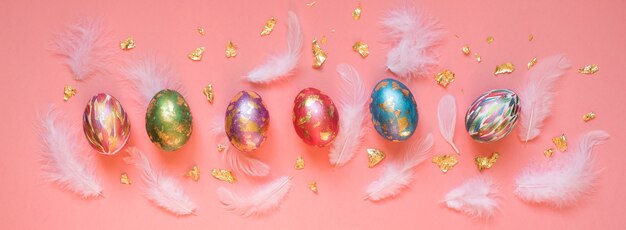 Oeufs de Pâques peints colorés avec des plumes blanches et des morceaux de feuille d'or sur le fond rose. Bannière de Pâques.