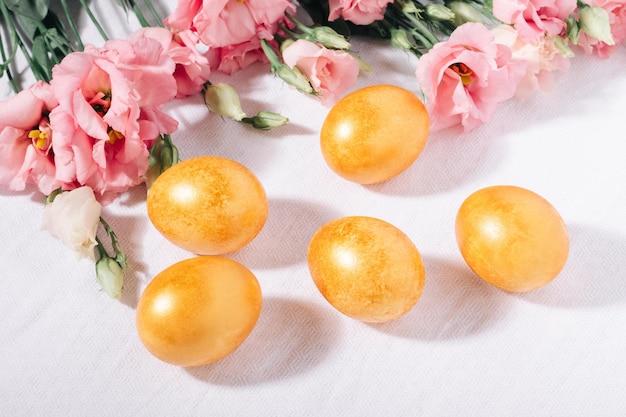 Oeufs de Pâques peints en or dans des tons pastel et fleurs roses sur une nappe en coton blanc