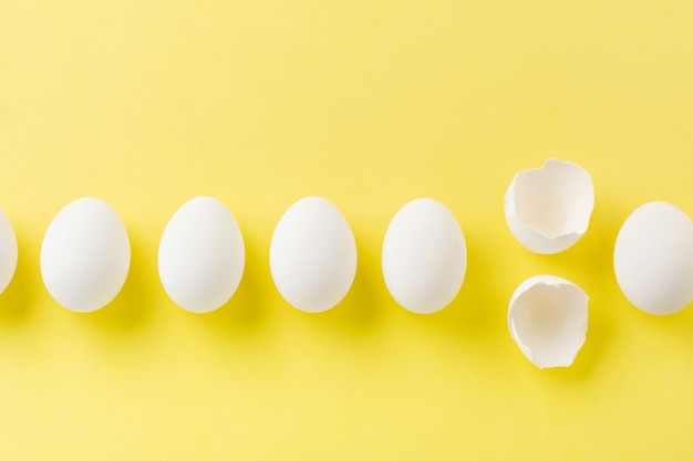 Oeufs de poule crus blancs couchés dans la rangée horizontale avec oeuf cassé sur une surface jaune. Vue de dessus.