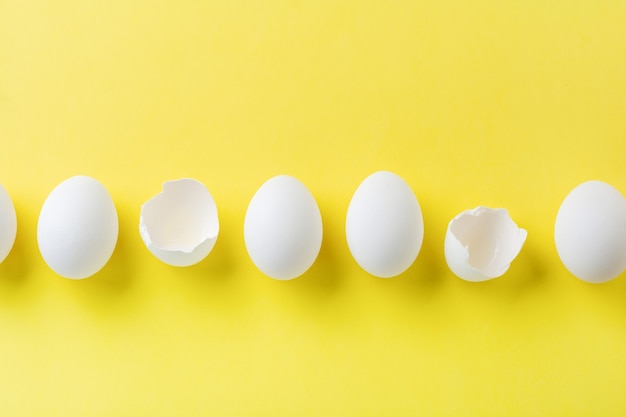 Oeufs de poule crus blancs couchés dans la rangée horizontale avec oeuf cassé sur une surface jaune. Vue de dessus.