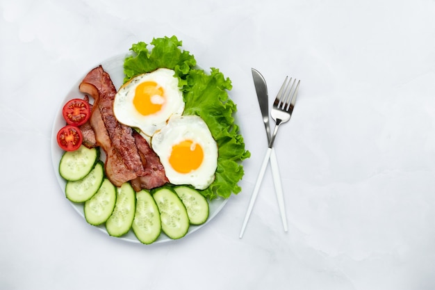 Oeufs de poule frits avec bacon et légumes sur une table grise. Notion de petit-déjeuner. Vue de dessus. Fond de nourriture le matin. Concombres et tomates frais tranchés. Composition à plat.