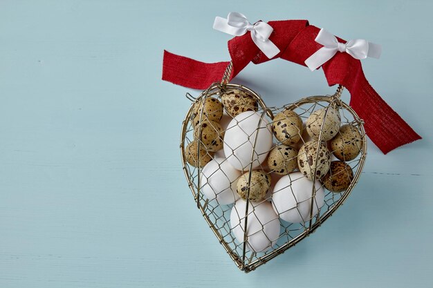 Oeufs de poulet et de caille dans une boîte en fil de métal tissé en forme de coeur attachée avec un ruban sur un fond bleu clair