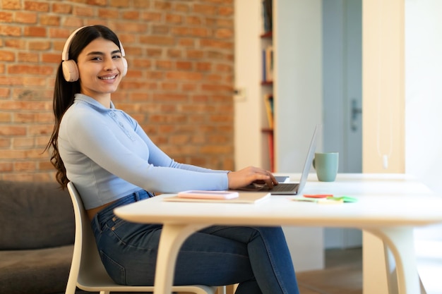 Offre d'apprentissage en ligne heureuse adolescente apprenant en ligne avec un ordinateur portable et des écouteurs assis au bureau à la maison