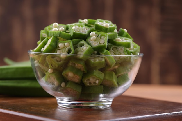 Okra ou Ladys finger ou Bhindi légumes verts frais disposés sur une planche de bois avec un bol en verre plein d'anneaux tranchés de gombo avec mise au point sélective sur fond de bois