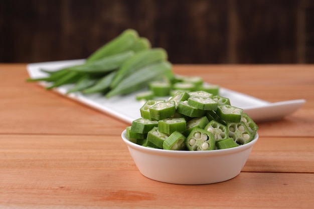 Okra ou Ladys finger ou Bhindi légumes verts frais disposés sur une vaisselle blanche avec un bol rempli d'anneaux tranchés de gombo avec mise au point sélective sur fond de bois