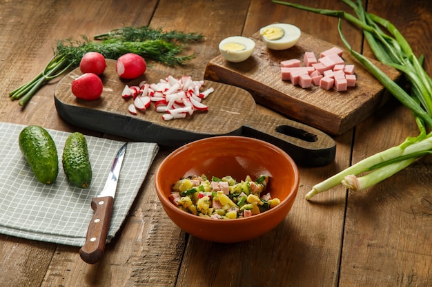Okroshka dans une assiette d'argile sur une table sur une table en bois à côté de légumes sur des planches et un couteau.
