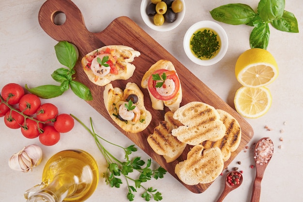 Olives, huile, crevettes grillées et salade de légumes frais sur une planche de bois sur une planche de pierre en ardoise noire sur une surface sombre, tomates juteuses sur du pain frais, pesto comme garniture. vue de dessus. Mise à plat