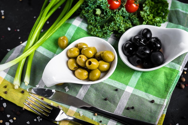 Olives et olives dans des bols blancs, légumes avec un chiffon et une planche de bois, couverts sur fond noir