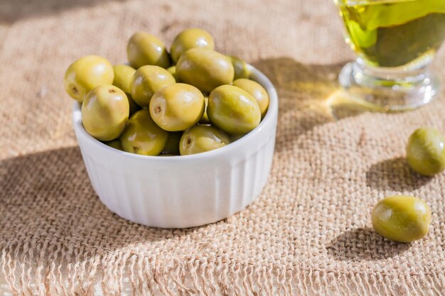 Photo olives vertes fraîches marinées dans un bol de céramique blanche et une sauce à l'huile d'olive vierge de qualité supérieure sur fond de tissu de burlap