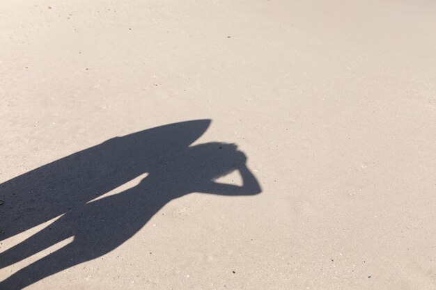 Une ombre d'une femme profitant du temps libre sur la plage par une journée ensoleillée, surfant, debout avec sa planche de surf, couvrant ses yeux du soleil.