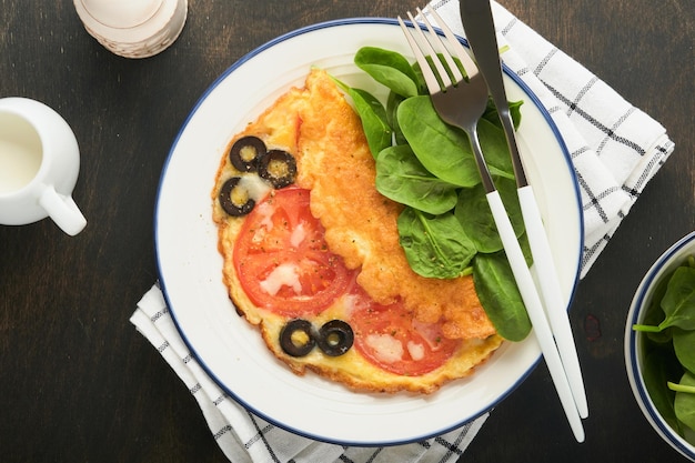 Omelette aux tomates, olives noires, fromage cottage et herbes d'épinards vertes sur une plaque blanche sur un vieux fond sombre rustique en bois Délicieux petit-déjeuner Nourriture de petit-déjeuner sain Vue de dessus