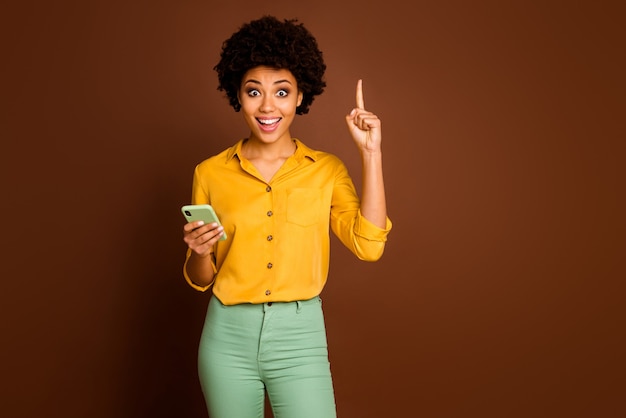 Omg je sais quel texte! Excité fou blogueur fille afro-américaine utiliser le type de smartphone sms pense obtenir une idée incroyable lever le doigt porter jaune élégant tenue verte couleur marron isolé