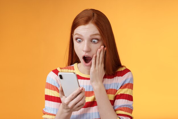 Omg quoi diable. Portrait choqué concerné jeune rousse sensible impressionné femme rousse regard smartphone écran tactile joue la mâchoire baisse étourdi surpris debout fond orange tenir le téléphone.