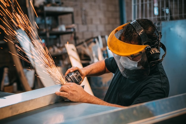 On voit un homme adulte travailler à couper un morceau de métal dans un atelier industriel. On voit des étincelles.