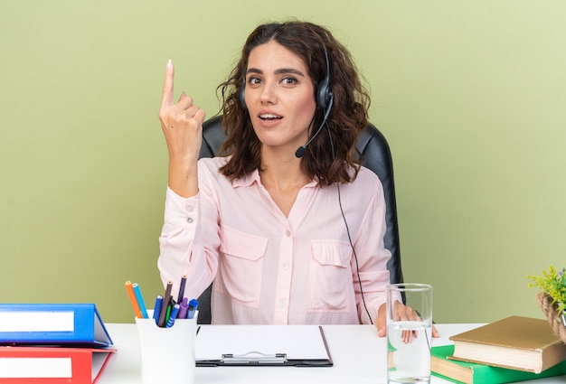 Opérateur de centre d'appels féminin assez surpris sur un casque assis au bureau avec des outils de bureau pointant vers le haut