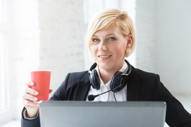 Opérateur de centre d'appels femme souriante vêtue d'un costume élégant noir avec écouteurs casque tenant un café pendant la pause