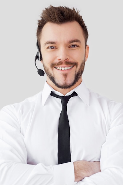 Opérateur masculin confiant. Beau jeune homme en tenue de soirée et casque regardant la caméra et souriant en se tenant debout sur fond gris