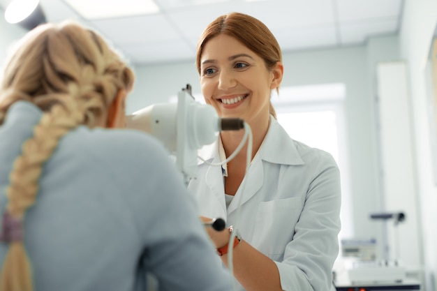 Ophtalmologiste joyeux. Ophtalmologiste roux gai souriant tout en s'asseyant devant la fille