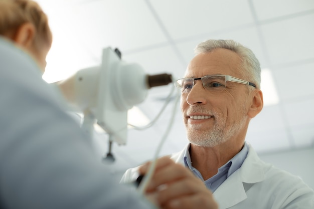 Photo ophtalmologiste souriant. ophtalmologiste aux cheveux gris barbu portant des lunettes et un uniforme blanc souriant