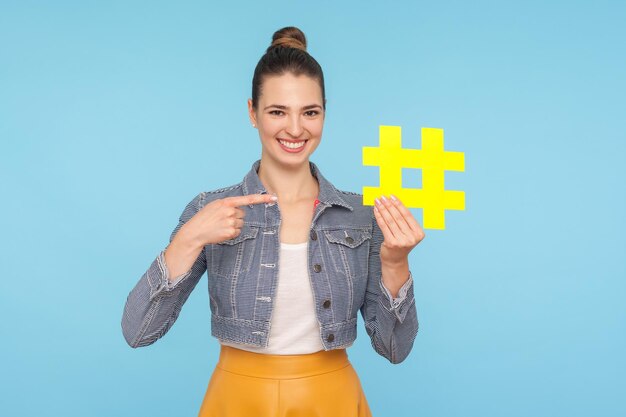 Optimiste heureuse jolie femme avec un chignon dans une tenue élégante pointant vers le grand symbole de hashtag jaune et souriant à la promotion de la caméra des tendances du blog tourné en studio intérieur isolé sur fond bleu