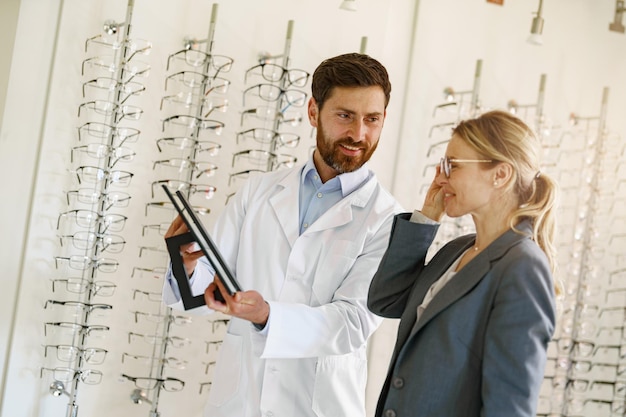 Optométriste souriant aidant une femme à choisir des lunettes dans un magasin d'optique montrant à son miroir