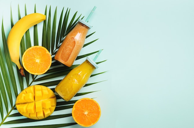 Orange fraîche, banane, ananas, smoothie à la mangue et fruits juteux sur feuilles de palmier sur fond bleu. Boisson d&#39;été détox.