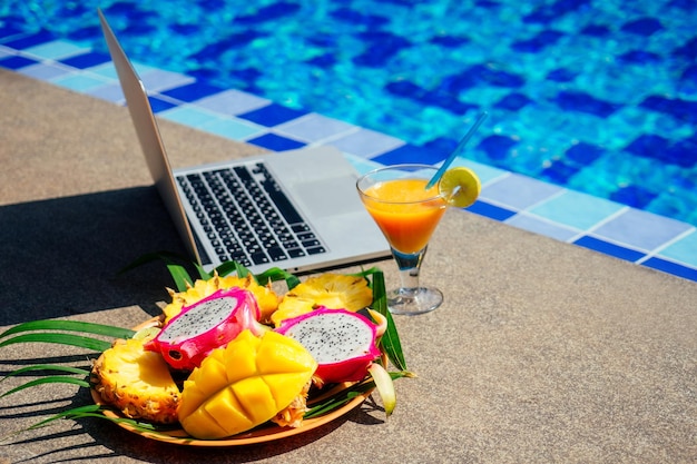 Ordinateur portable à coups de fruits tropicaux frais papaye, ananas, fruit du dragon au bord de la piscine travail à distance indépendant et frilansing.