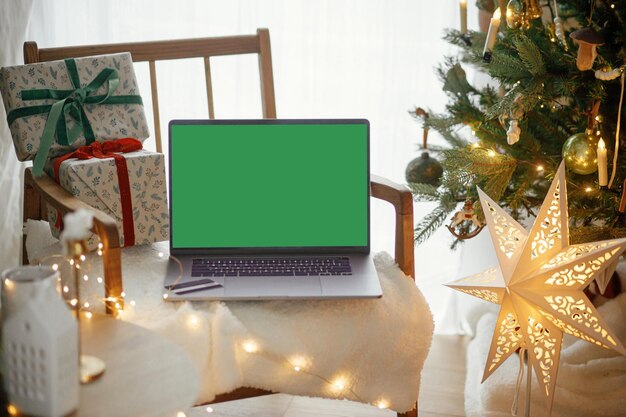 Ordinateur portable avec écran vide et cadeaux de Noël élégants sur une chaise moderne sur fond d'arbres décorés de fête lumières dorées et étoile dans la chambre achats de Noël en ligne et ventes maquette d'ordinateur portable