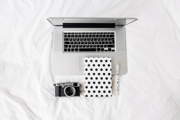 Ordinateur portable, écriture de notes et appareil photo sur lit blanc travail à la maison concept. Vue de dessus d'un bureau de travail.
