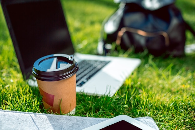 Ordinateur portable sur l'herbe verte avec tasse à café, sac et tablette dans un parc extérieur. Espace de copie. Nature morte