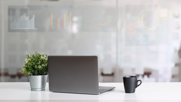 Ordinateur portable mettant sur une table blanche moderne avec une tasse à café en céramique, plante en pot avec des graphiques et fond de signal de bougie dans le salon.