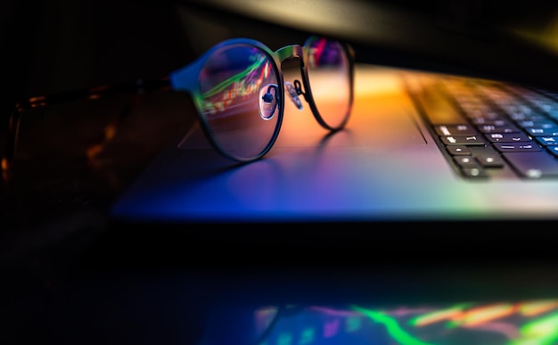 Un ordinateur portable à moitié fermé dans le noir avec une lueur colorée et des lunettes