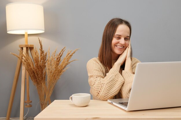 Ordinateur portable pour le travail à distance Attractive jolie femme aux cheveux bruns portant un pull beige travaillant sur l'ordinateur en regardant l'écran avec un sourire joyeux tout en étant assise à table contre la lampe et les fleurs sèches