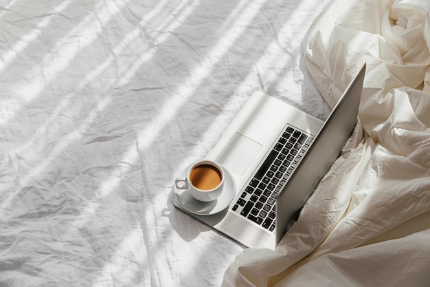 Ordinateur portable et tasse de café sur un lit blanc avec une couverture. Concept de travail à domicile. Lumière du matin. Concept de mode de vie