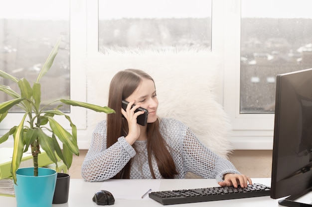 Organisation du bureau à domicile, dispositif de travail à distance. Une jeune fille est assise à une table, tape sur un ordinateur, analyse des documents, sourit, parle au téléphone